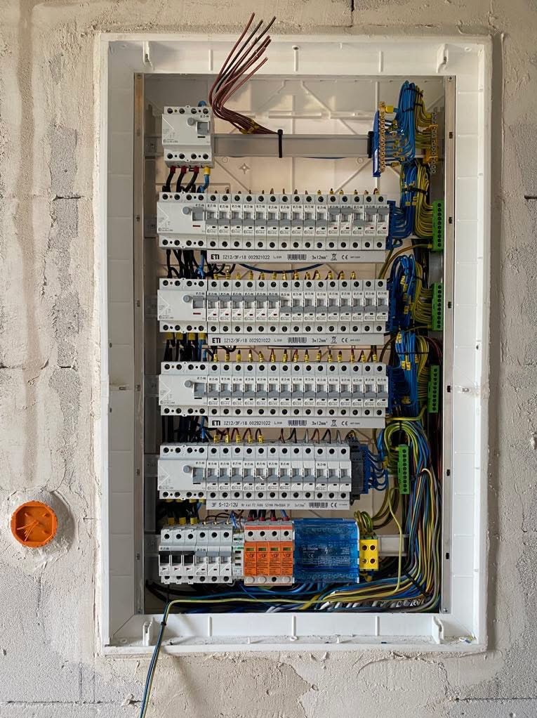 Rozdzielnia elektryczna z licznymi zabezpieczeniami i okablowaniem, zamontowana w surowej ścianie. Widoczna pomarańczowa puszka instalacyjna po lewej stronie.