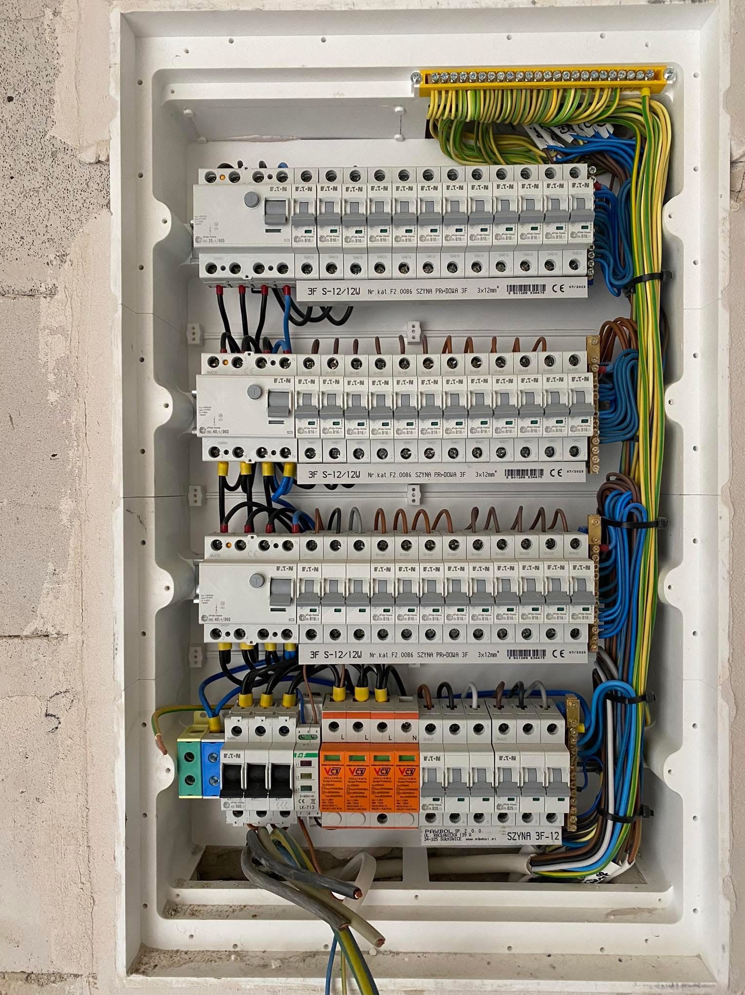 Rozdzielnica elektryczna w białej obudowie, widoczne zabezpieczenia, przewody w różnych kolorach, szyny. Surowa ściana w tle.
