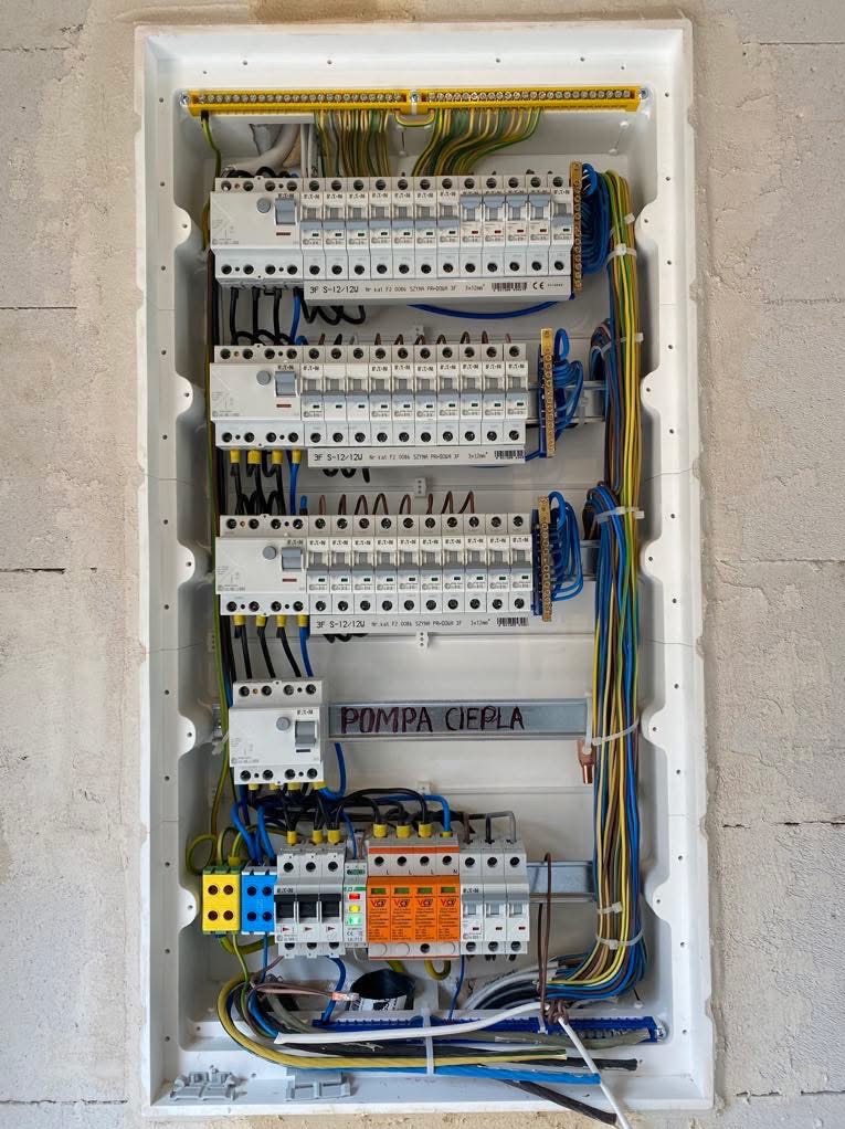 Rozdzielnica elektryczna w białej obudowie, widoczne bezpieczniki, kable w różnych kolorach, szyna z napisem 'POMPA CIEPŁA'.
