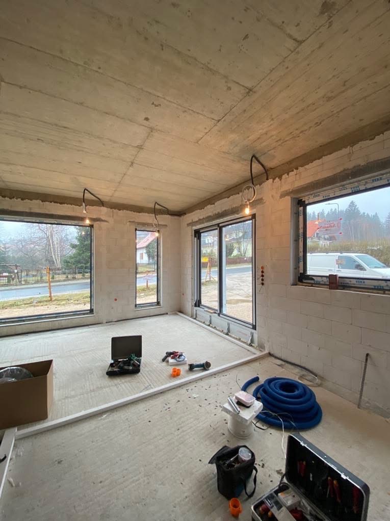 Surowy stan wnętrza z widocznymi przewodami oświetleniowymi, narzędziami i materiałami budowlanymi. Ściany z betonu komórkowego, duże okna z widokiem na zewnątrz.