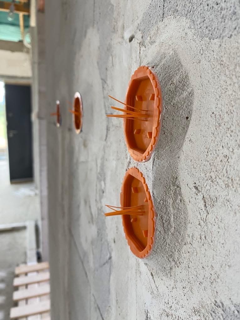 Ściana w trakcie prac wykończeniowych z zamontowanymi, pomarańczowymi puszkami elektrycznymi pod gniazdka. Widoczne przewody. Surowy tynk.