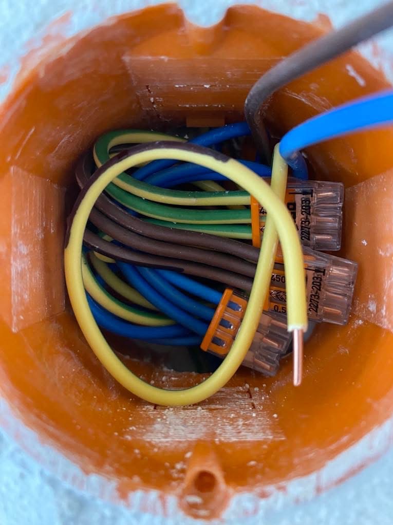 Pomarańczowa puszka instalacyjna z widocznymi przewodami w różnych kolorach (niebieski, brązowy, żółto-zielony) i złączkami Wago. Widok z góry.