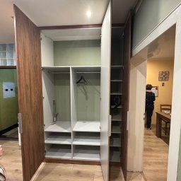 MM Bespoke interiors - meble na wymiar - Otwarta szafa wnękowa z drewnianymi drzwiami i białym wnętrzem, widoczne półki i drążek na wieszaki. Dziewczynka przygląda się szafie. W tle widoczne inne pomieszczenie.