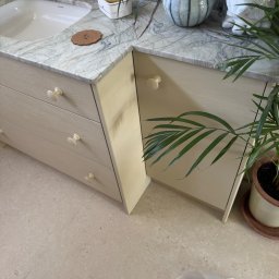 MM Bespoke interiors - meble na wymiar - Jasna szafka łazienkowa z szufladami i marmurowym blatem, na którym stoją dekoracje i akcesoria. Uwagę przykuwają kremowe gałki szuflad i doniczka z rośliną.