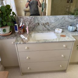 MM Bespoke interiors - meble na wymiar - Łazienka z szafką pod umywalką z trzema szufladami, marmurowym blatem i roślinami. W lustrze odbija się osoba robiąca zdjęcie.