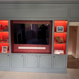 MM Bespoke interiors - meble na wymiar - Niebieska zabudowa meblowa z podświetlanymi na czerwono półkami, wnęką na TV i szafkami. Dekoracje na półkach, widoczny fragment podłogi i przejście do innego pomieszczenia.