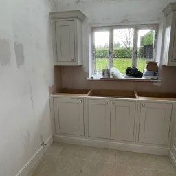 MM Bespoke interiors - meble na wymiar - Montaż szafek kuchennych w Bielsko-Białej. Kremowe fronty z uchwytami, widoczne puszki i akcesoria na parapecie. Ściany przygotowane do malowania.