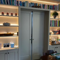 Biała zabudowa biblioteczna z podświetlanymi półkami, na których stoją książki, dekoracje i naczynia. W centralnej części przesuwne drzwi. Widok na wnętrze.