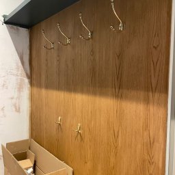 MM Bespoke interiors - meble na wymiar - Panel ścienny z drewnopodobną okleiną i złotymi haczykami, zamontowany w przedpokoju. Widoczne kartony i fragmenty ściany w trakcie wykończenia.