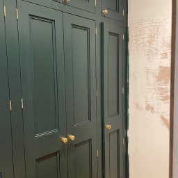 MM Bespoke interiors - meble na wymiar - Ciemnozielona szafa wnękowa z frezowanymi drzwiami i złotymi gałkami na tle ściany w trakcie malowania. Widoczne niedoskonałości wykończenia.