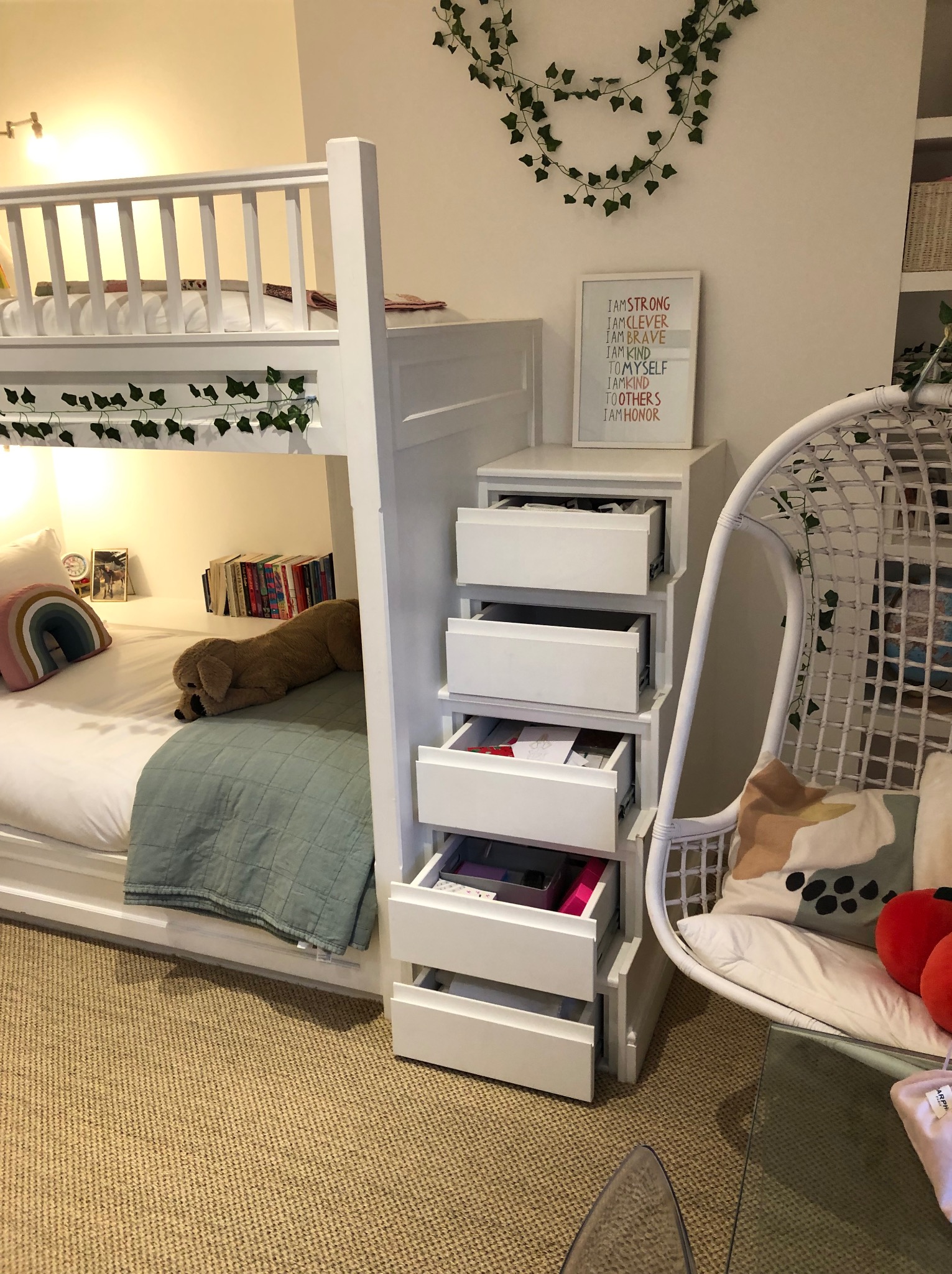 Białe łóżko piętrowe z wbudowanymi szufladami, ozdobione girlandą z bluszczu. W tle białe ściany, wiszące krzesło i półki z koszami. Przytulny pokój dziecięcy.