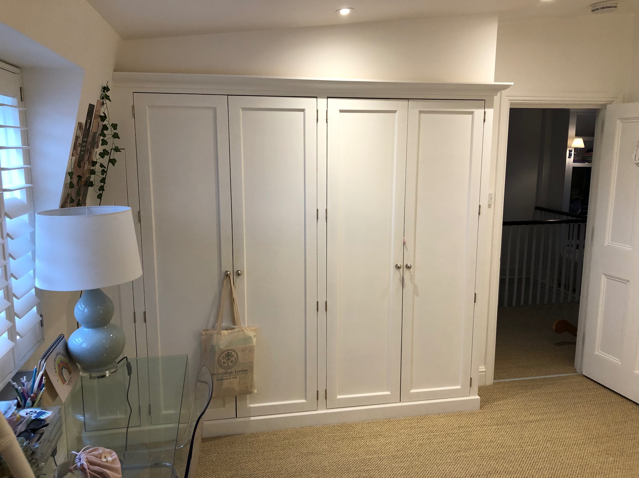 Biała szafa wnękowa z czterema drzwiami, z torbą materiałową wiszącą na jednych drzwiach, w jasnym pokoju z otwartymi drzwiami do innego pomieszczenia.