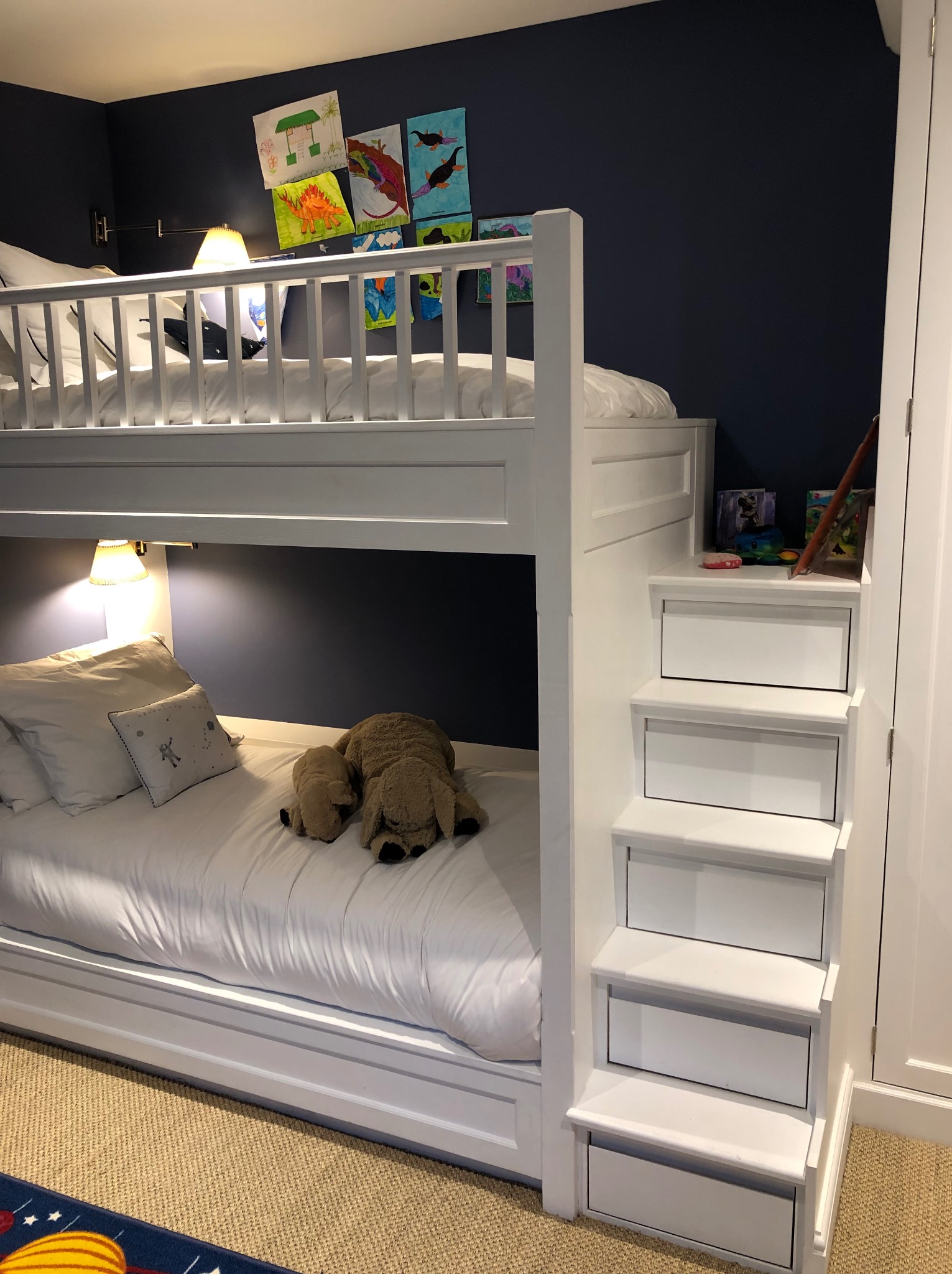 Białe łóżko piętrowe z drabinką-schowkiem w pokoju dziecięcym. Na dolnym łóżku pluszowe zabawki, na ścianie rysunki dziecięce. Ciemna ściana, lampki nocne.