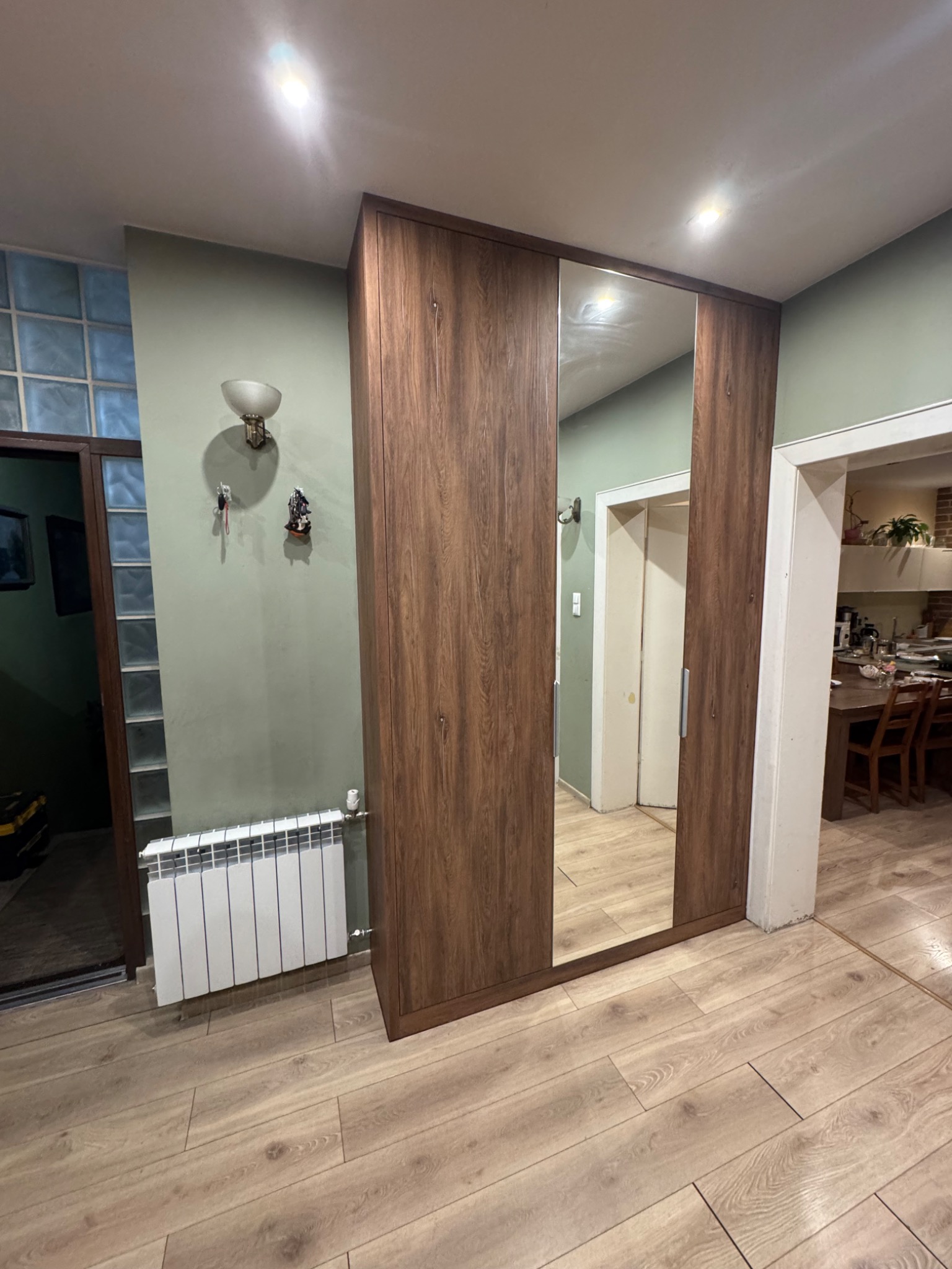 Szafa wnękowa z lustrem w brązowej okleinie, wkomponowana w przedpokój z drewnianą podłogą i grzejnikiem. Widok na wnętrze mieszkania.