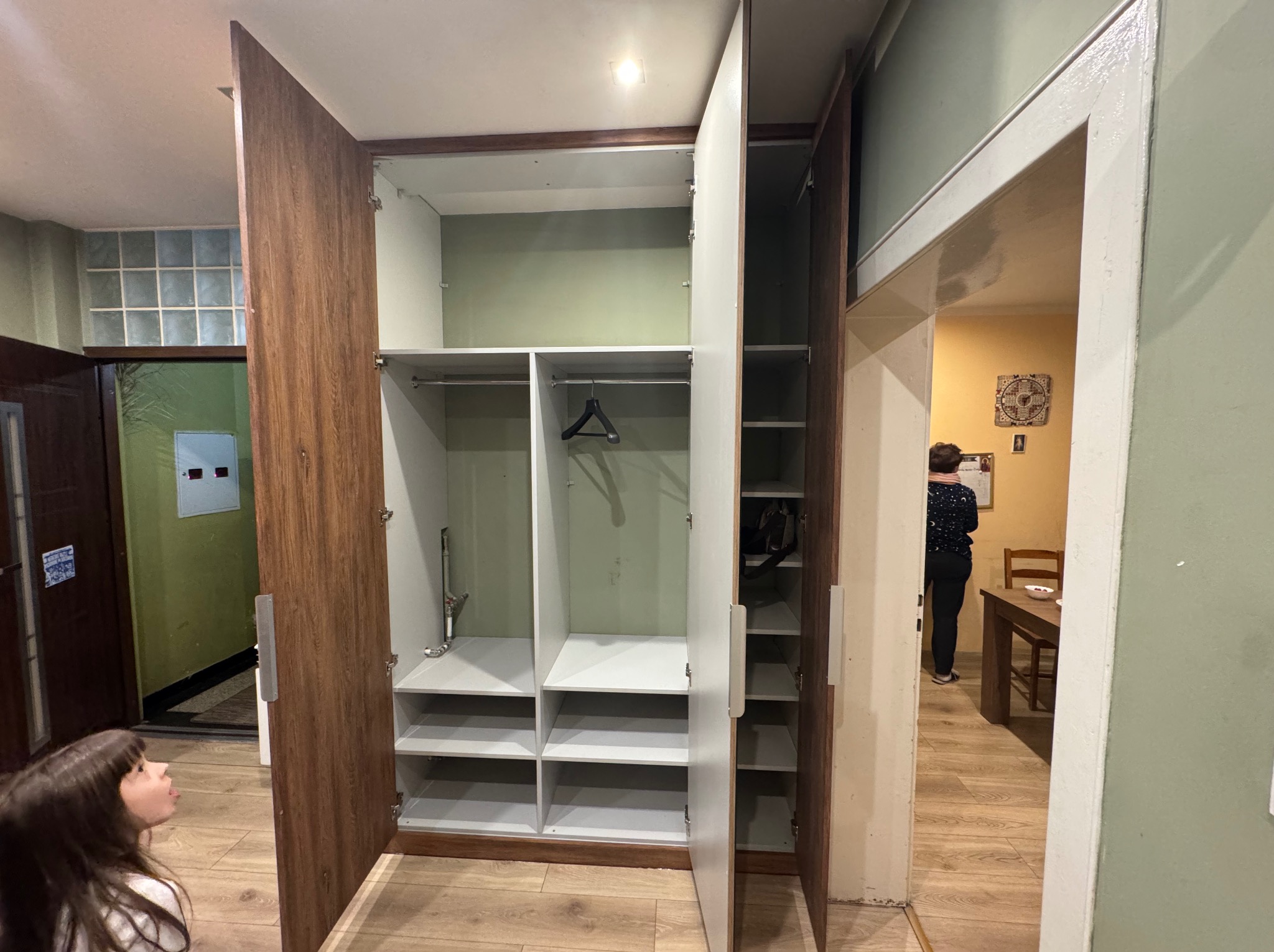 Otwarta szafa wnękowa z drewnianymi drzwiami i białym wnętrzem, widoczne półki i drążek na wieszaki. Dziewczynka przygląda się szafie. W tle widoczne inne pomieszczenie.