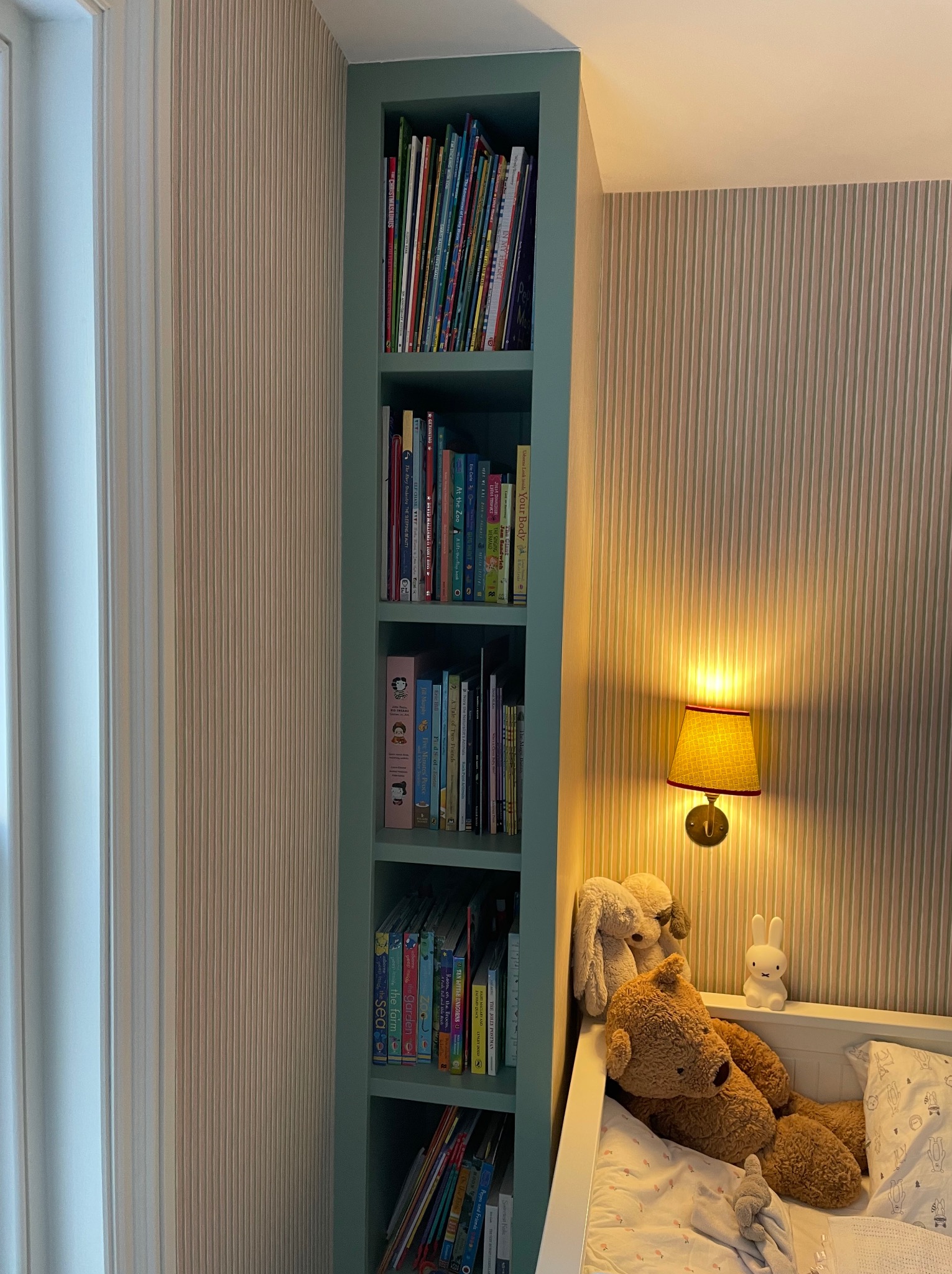 Wąska, wnękowa szafa na książki w pokoju dziecięcym, pomalowana na pastelowy zielony kolor. Obok łóżeczko z pluszakami, a nad nim lampka nocna.