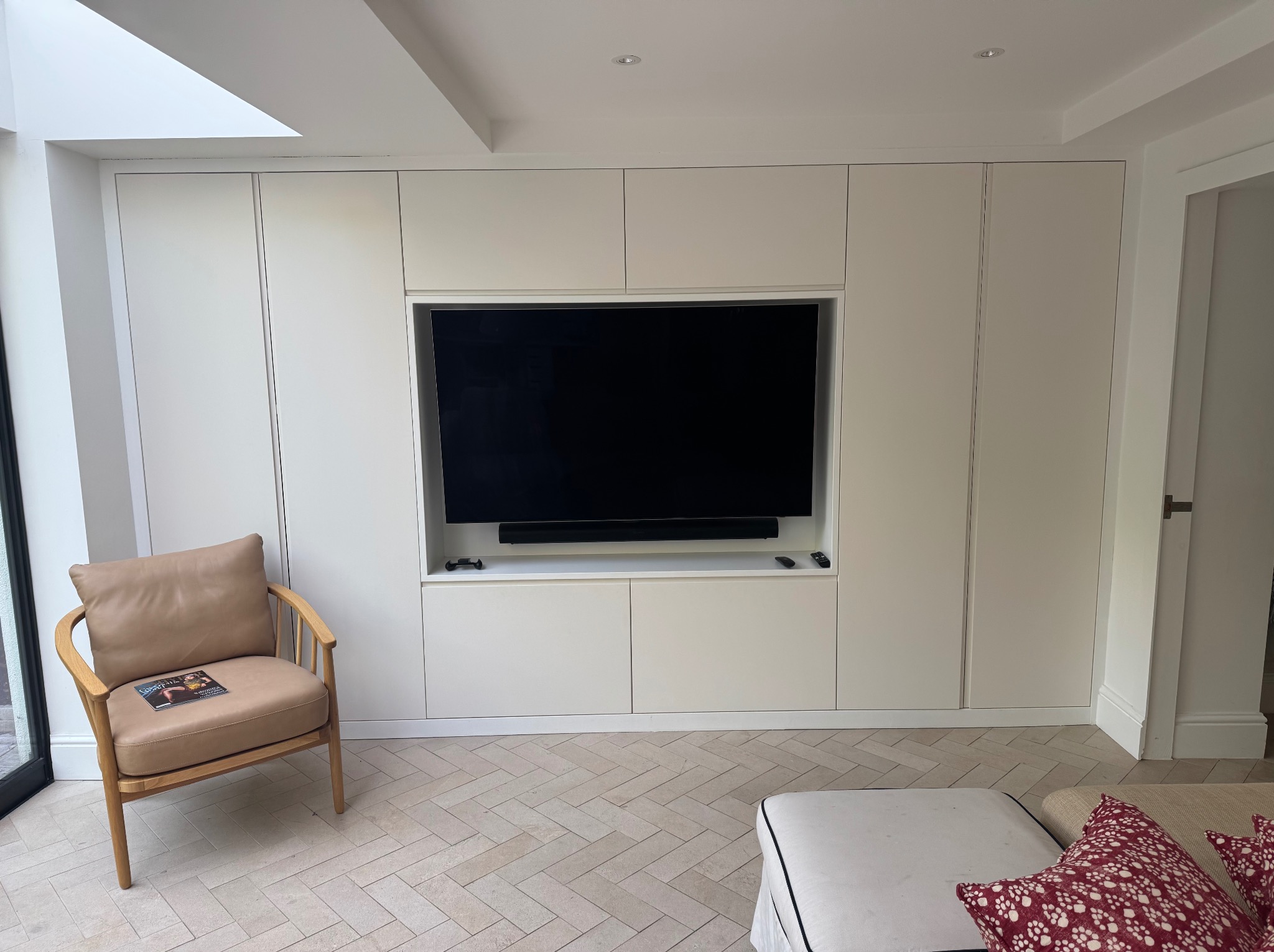 Biała szafa wnękowa z miejscem na telewizor, wkomponowana w ścianę. Na pierwszym planie fotel i fragment sofy. Parkiet ułożony w jodełkę.