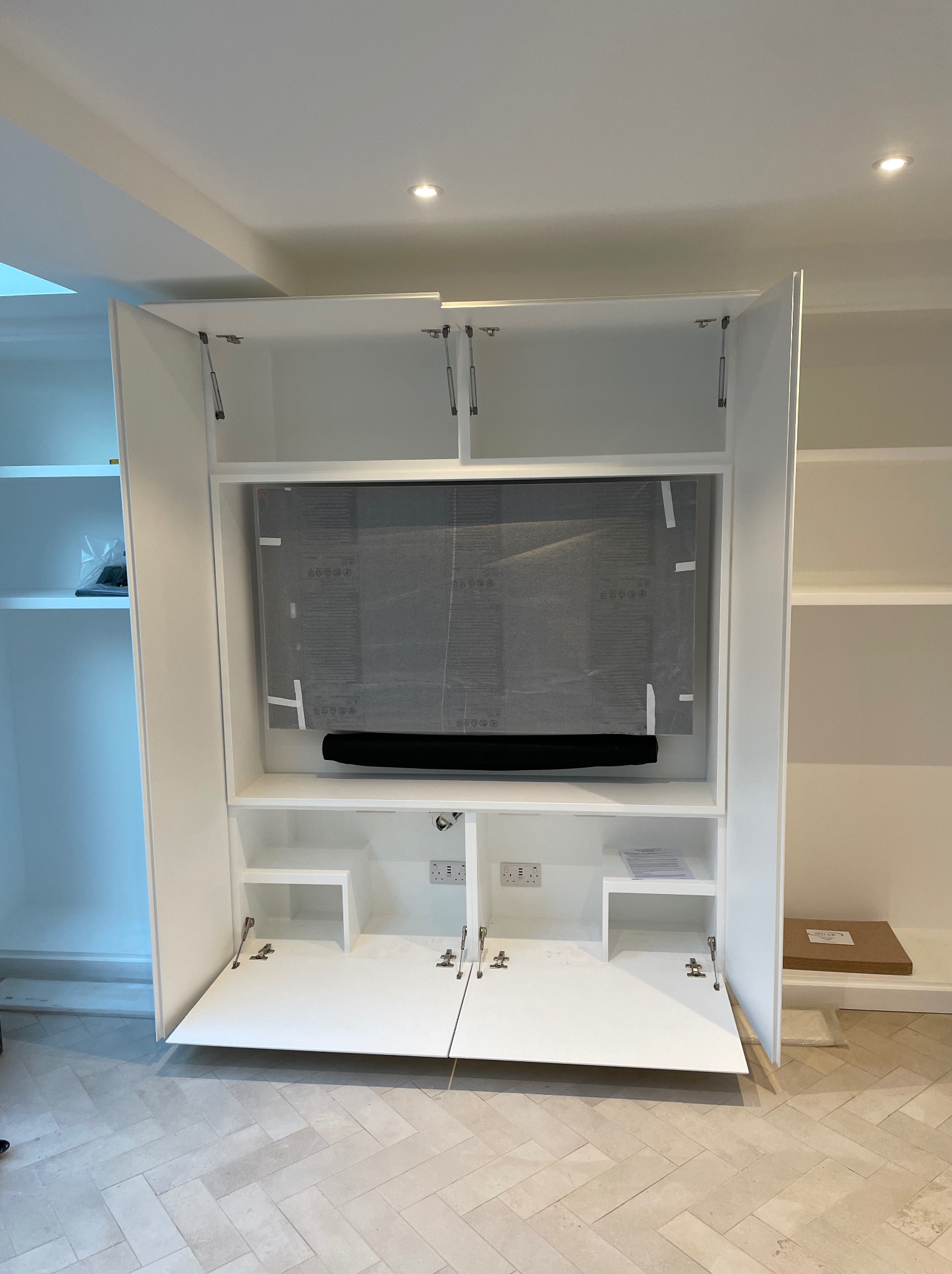Biała szafa wnękowa z otwartymi drzwiami, wewnątrz telewizor zabezpieczony folią. Widoczne gniazdka elektryczne i półki. Podłoga wyłożona jasną płytką w jodełkę.