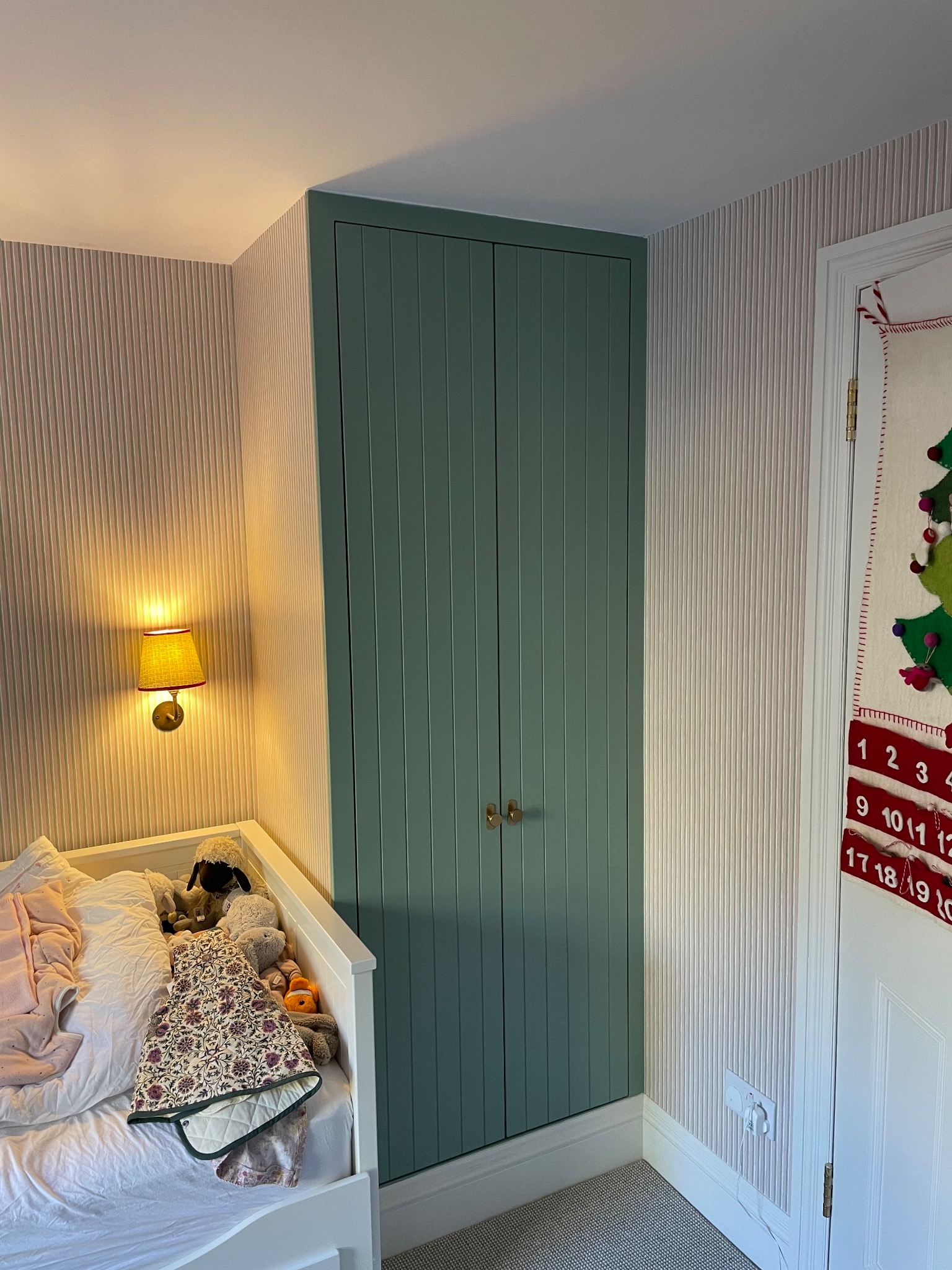 Zielona szafa wnękowa z drewnianymi uchwytami, w pokoju dziecięcym z tapetą w paski i łóżkiem z zabawkami. Oświetlenie boczne podkreśla fakturę szafy.