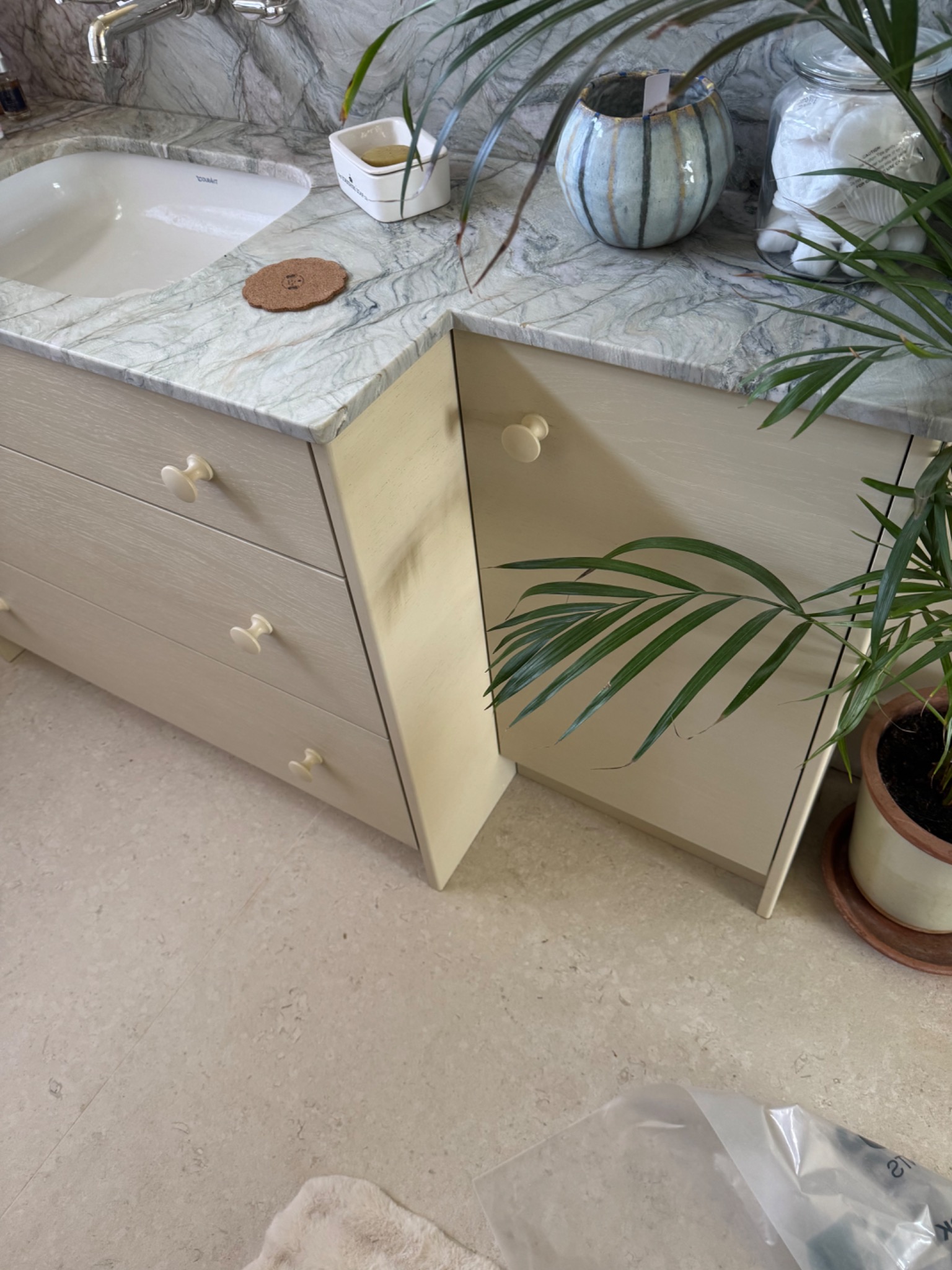 Jasna szafka łazienkowa z szufladami i marmurowym blatem, na którym stoją dekoracje i akcesoria. Uwagę przykuwają kremowe gałki szuflad i doniczka z rośliną.