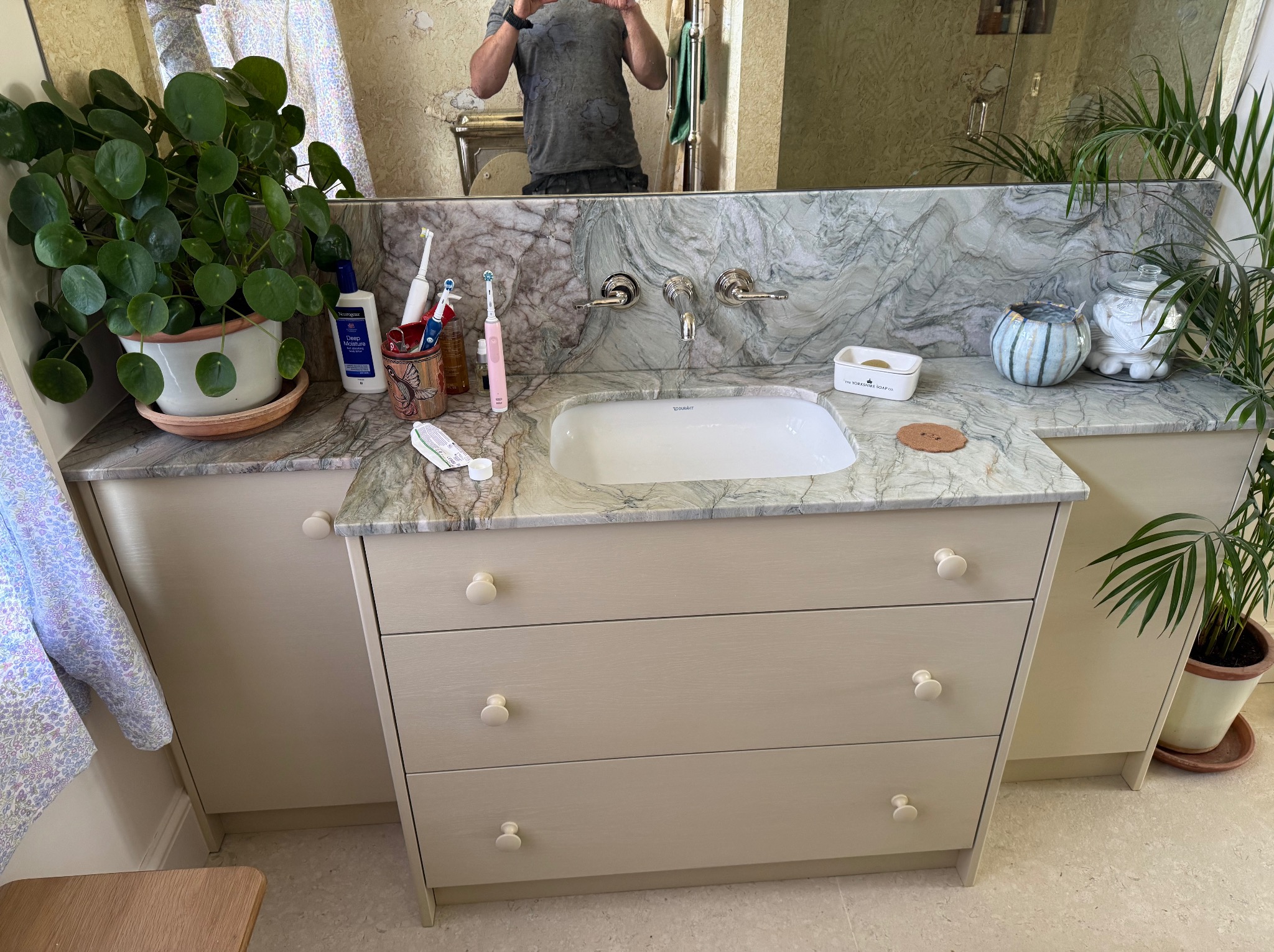 Łazienka z szafką pod umywalką z trzema szufladami, marmurowym blatem i roślinami. W lustrze odbija się osoba robiąca zdjęcie.