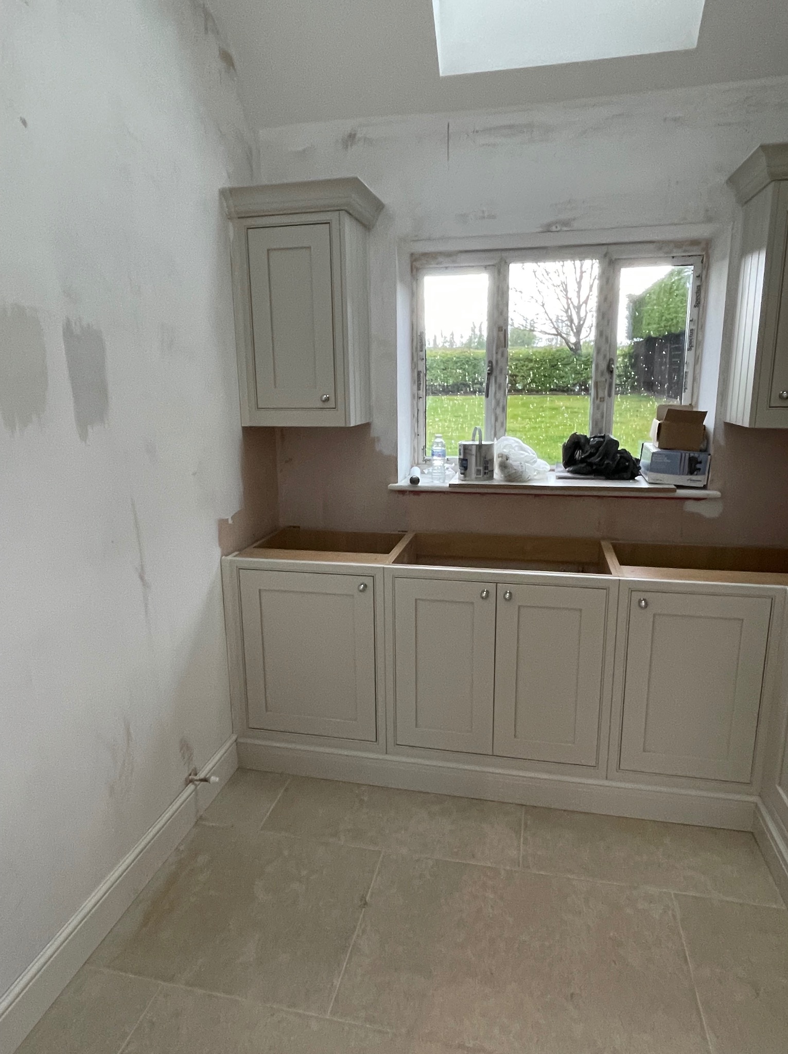 Montaż szafek kuchennych w Bielsko-Białej. Kremowe fronty z uchwytami, widoczne puszki i akcesoria na parapecie. Ściany przygotowane do malowania.