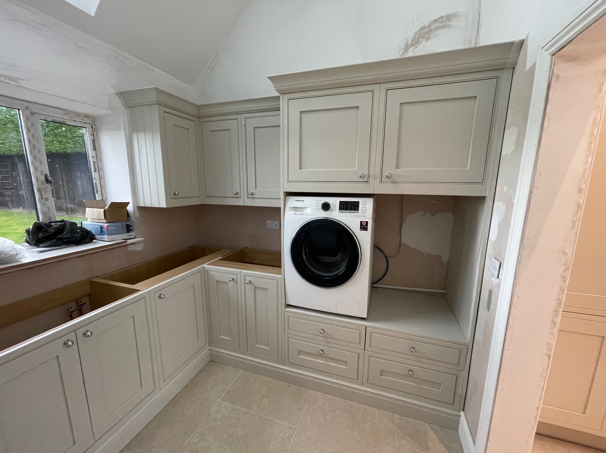 Jasna zabudowa meblowa z pralką Samsung wkomponowaną w szafki. Widoczne niedokończone prace wykończeniowe ścian. Ujęcie z perspektywy pod lekkim kątem.