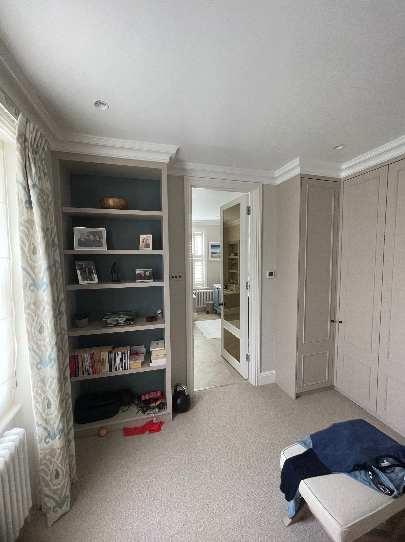 Jasne wnętrze z zabudową: szafa wnękowa z frezowanymi frontami i regał z dekoracjami. Widok na sąsiednie pomieszczenie przez otwarte drzwi.