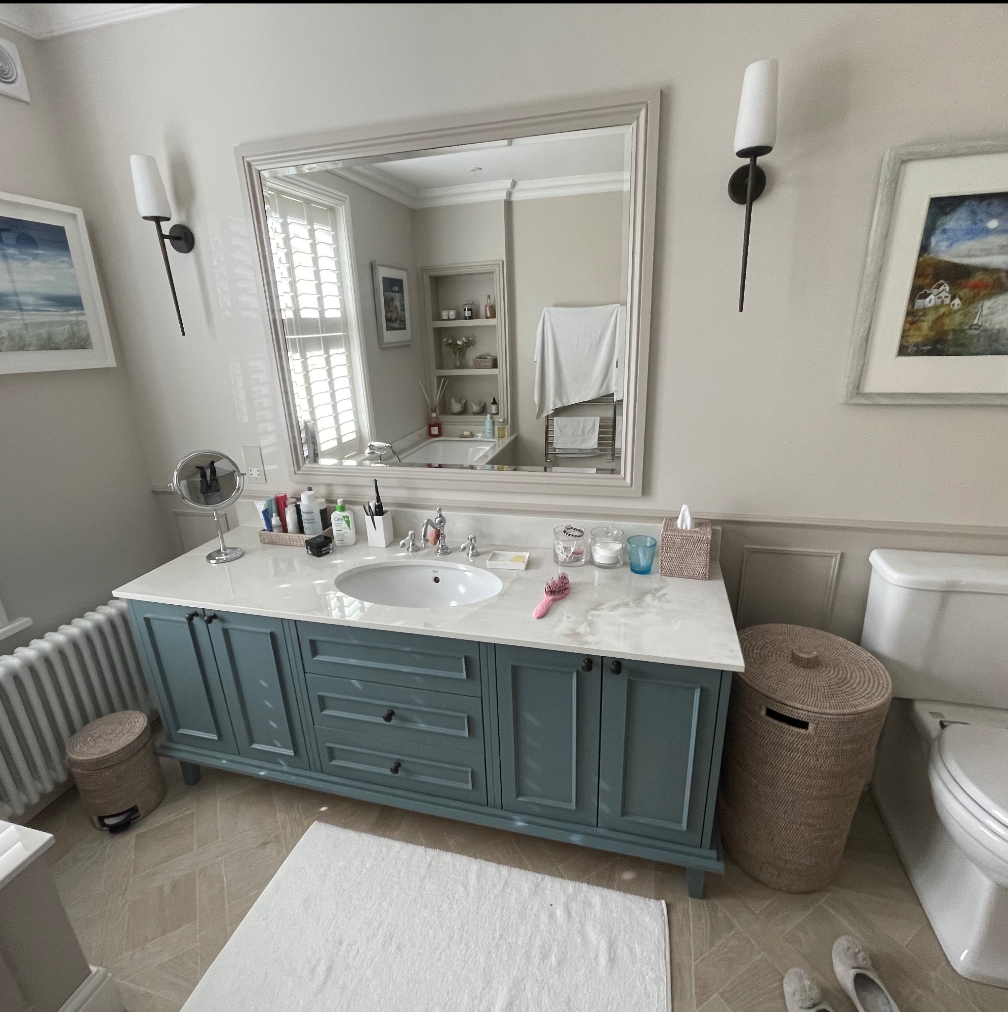 Niebieska szafka łazienkowa z marmurowym blatem, lustro w ramie, dekoracje i oświetlenie. Widok na wnętrze łazienki odbijający się w lustrze.