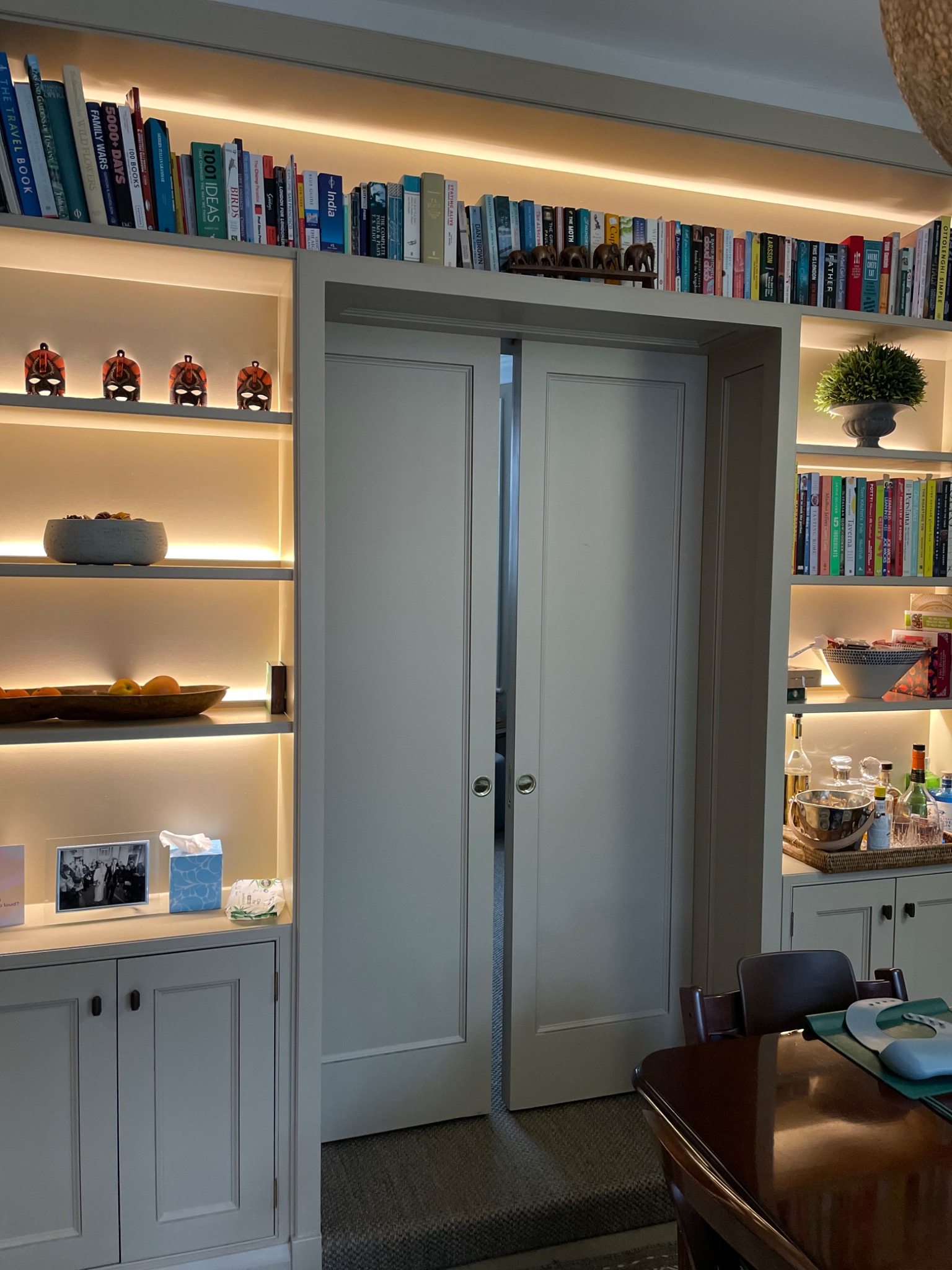 Biała zabudowa biblioteczna z podświetlanymi półkami, na których stoją książki, dekoracje i naczynia. W centralnej części przesuwne drzwi. Widok na wnętrze.