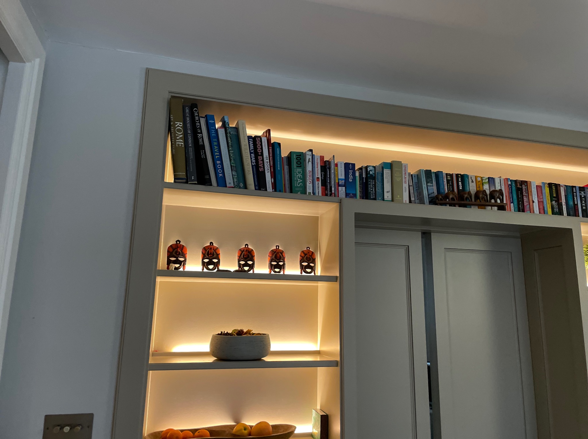 Biała szafa wnękowa z przesuwnymi drzwiami, podświetlonymi półkami wypełnionymi książkami, maskami i dekoracyjnymi misami. Ciepłe światło podkreśla detale.