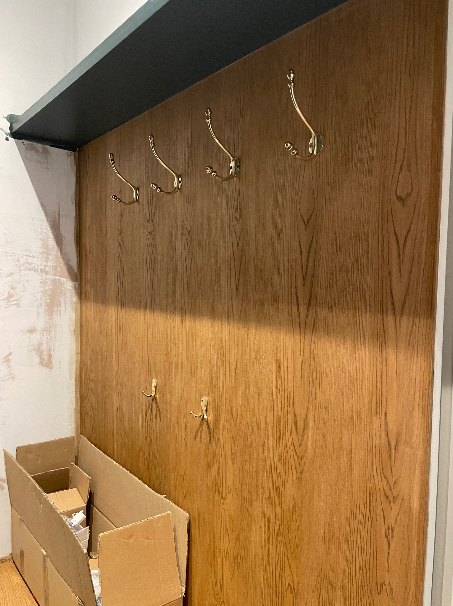 Panel ścienny z drewnopodobną okleiną i złotymi haczykami, zamontowany w przedpokoju. Widoczne kartony i fragmenty ściany w trakcie wykończenia.