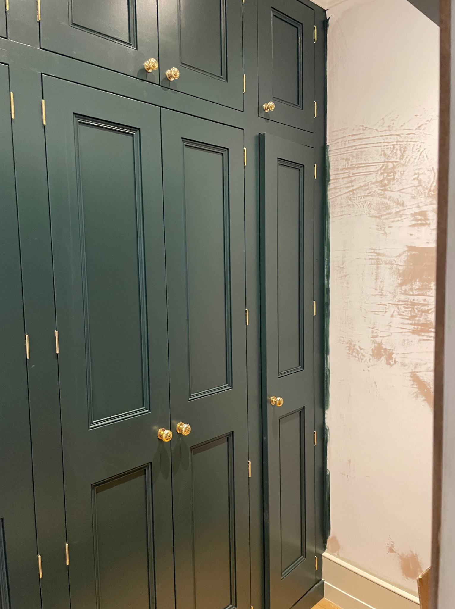 Ciemnozielona szafa wnękowa z frezowanymi drzwiami i złotymi gałkami na tle ściany w trakcie malowania. Widoczne niedoskonałości wykończenia.