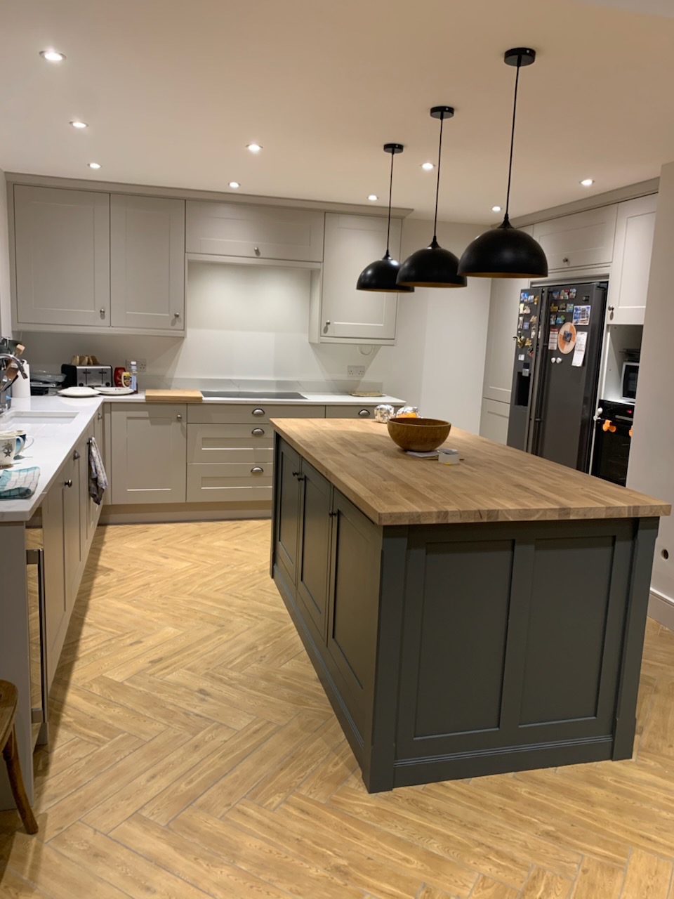 Jasna kuchnia z szarymi szafkami i wyspą z drewnianym blatem, oświetlona trzema czarnymi lampami wiszącymi. Parkiet ułożony w jodełkę.