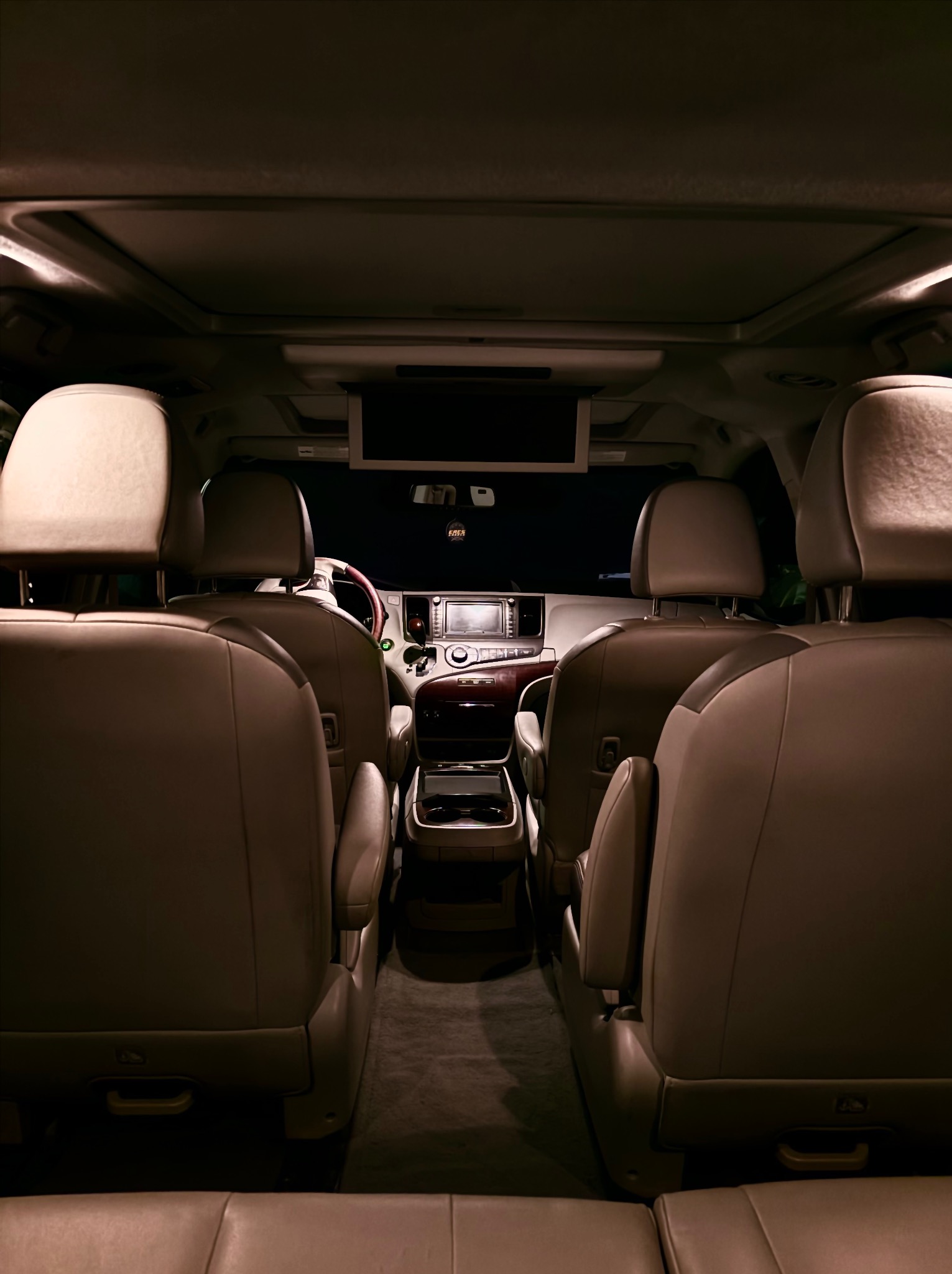 Wnętrze przestronnego busa z beżową skórzaną tapicerką, widok z tyłu na fotele, zagłówki i kokpit z elementami drewna. Oświetlenie wnętrza tworzy ciepłą atmosferę.