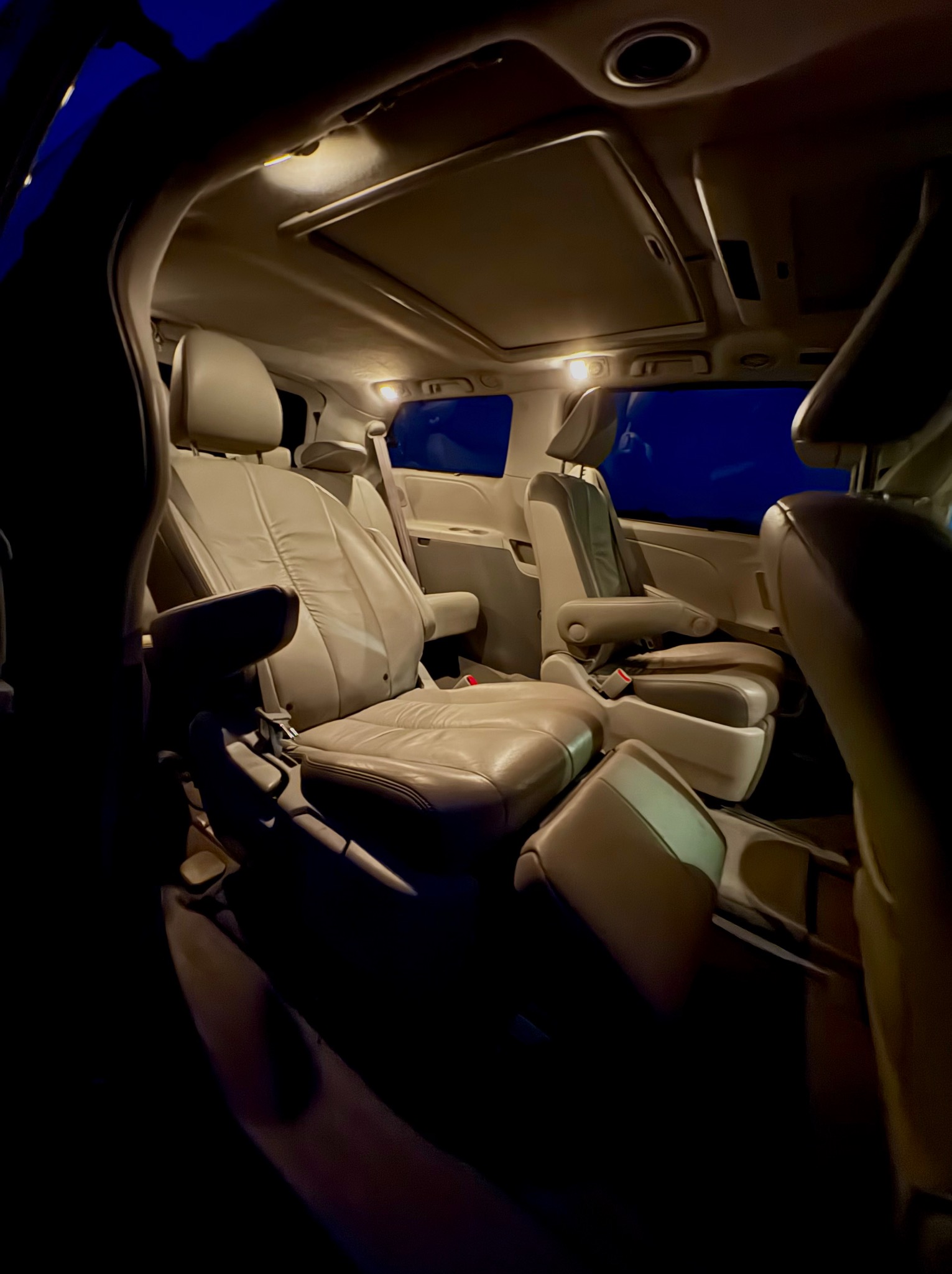 Wnętrze beżowego busa z rozsuwanymi drzwiami i skórzanymi fotelami, oświetlone lampkami sufitowymi, widok z perspektywy pasażera.