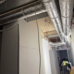 KOVALVENT SPÓŁKA Z OGRANICZONĄ ODPOWIEDZIALNOŚCIĄ - Montaż izolowanych przewodów wentylacyjnych pod sufitem, widoczne kable i fragment pracownika w kamizelce odblaskowej. Prace instalacyjne w budynku.