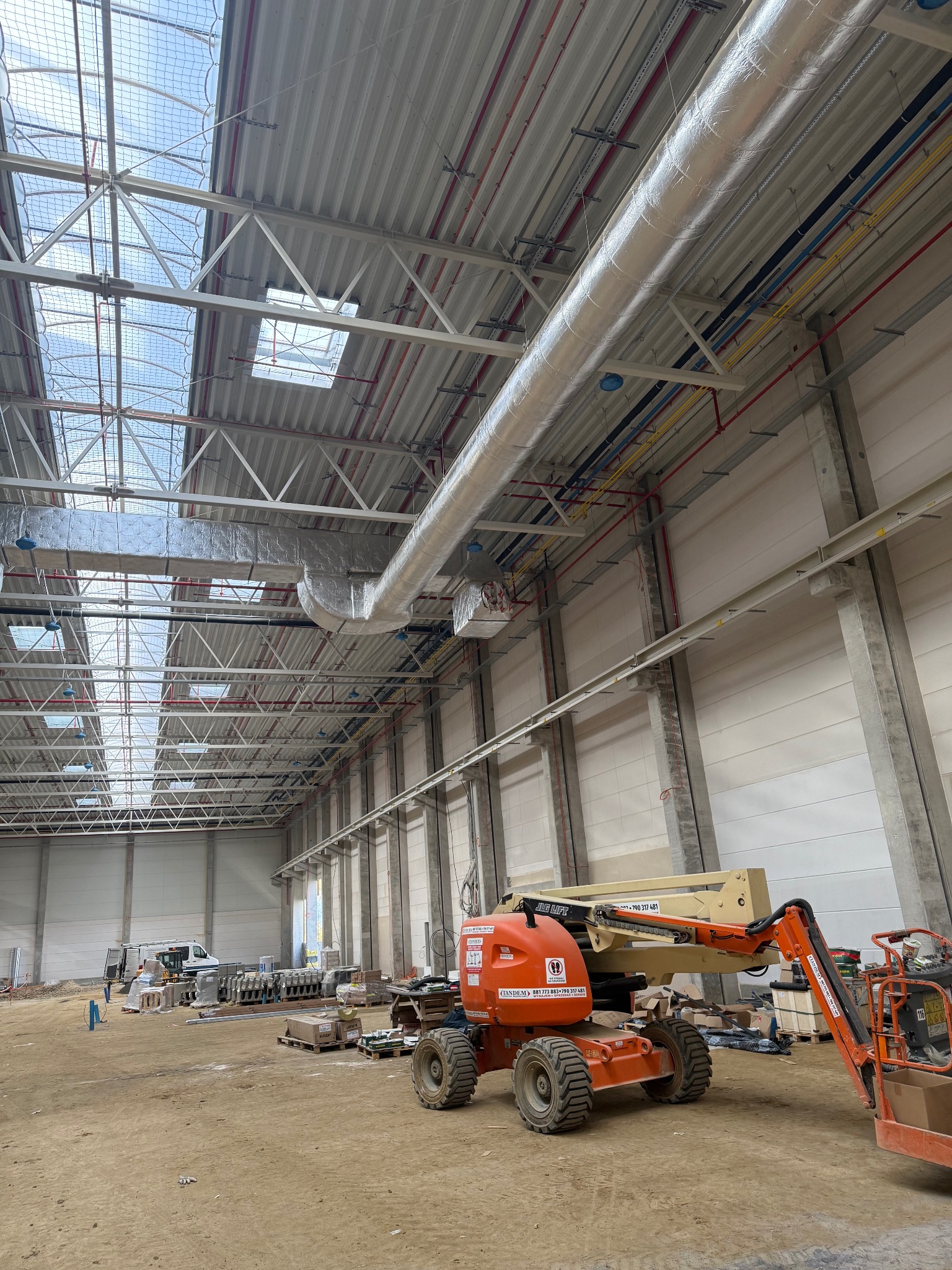 Montaż instalacji wentylacyjnej w hali przemysłowej. Widoczne kanały wentylacyjne pod sufitem, podnośniki koszowe i elementy konstrukcyjne budynku.