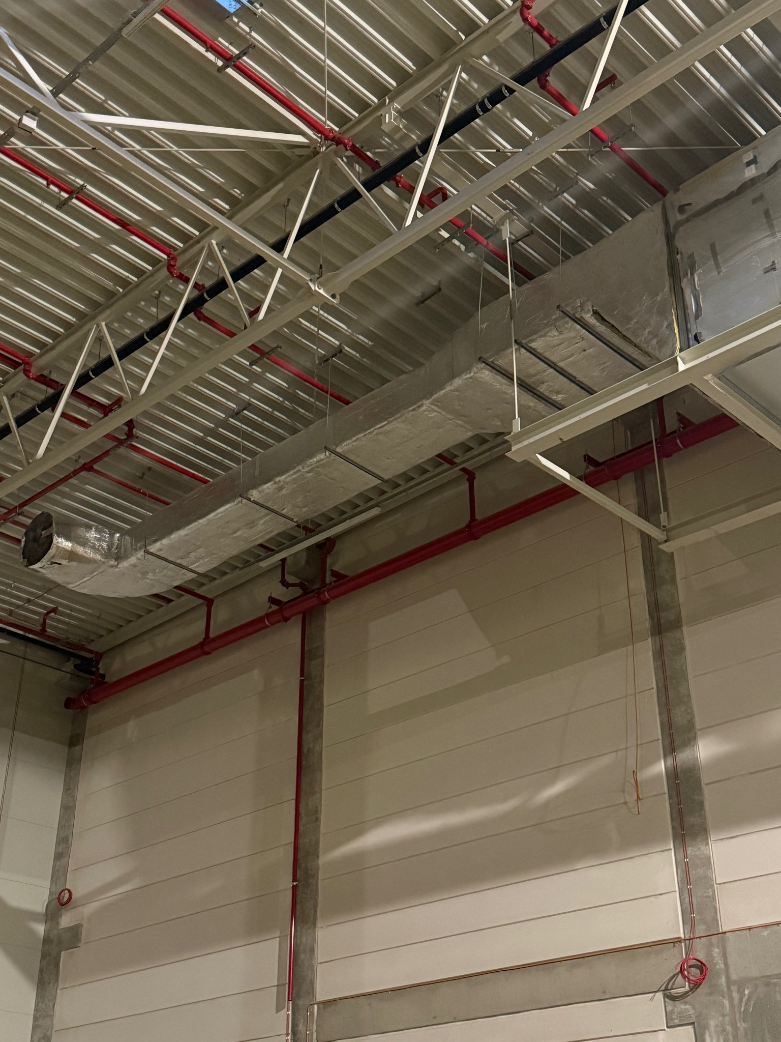 Szara, prostokątna instalacja wentylacyjna pod sufitem hali przemysłowej, z widocznymi czerwonymi rurami i metalową konstrukcją dachu. Ujęcie z dołu.