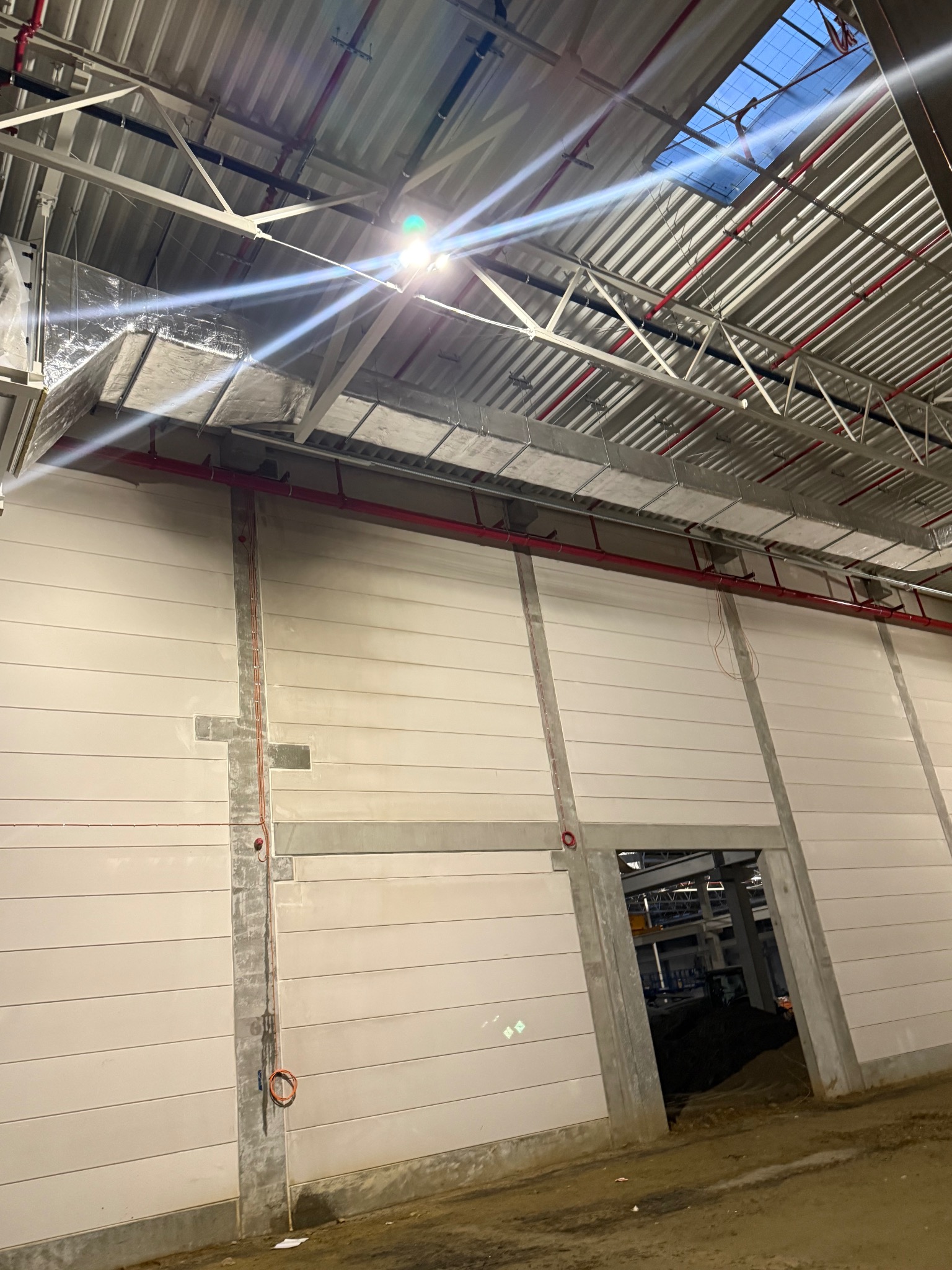 Wnętrze hali w trakcie budowy z widoczną instalacją wentylacyjną pod sufitem i czerwoną rurą. Widoczny fragment ściany z betonowymi elementami konstrukcyjnymi.