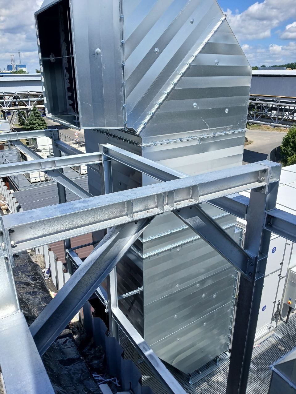 Srebrna, przemysłowa instalacja wentylacyjna na dachu budynku, widoczna z góry, z elementami konstrukcji wsporczej i fragmentami innych instalacji w tle.