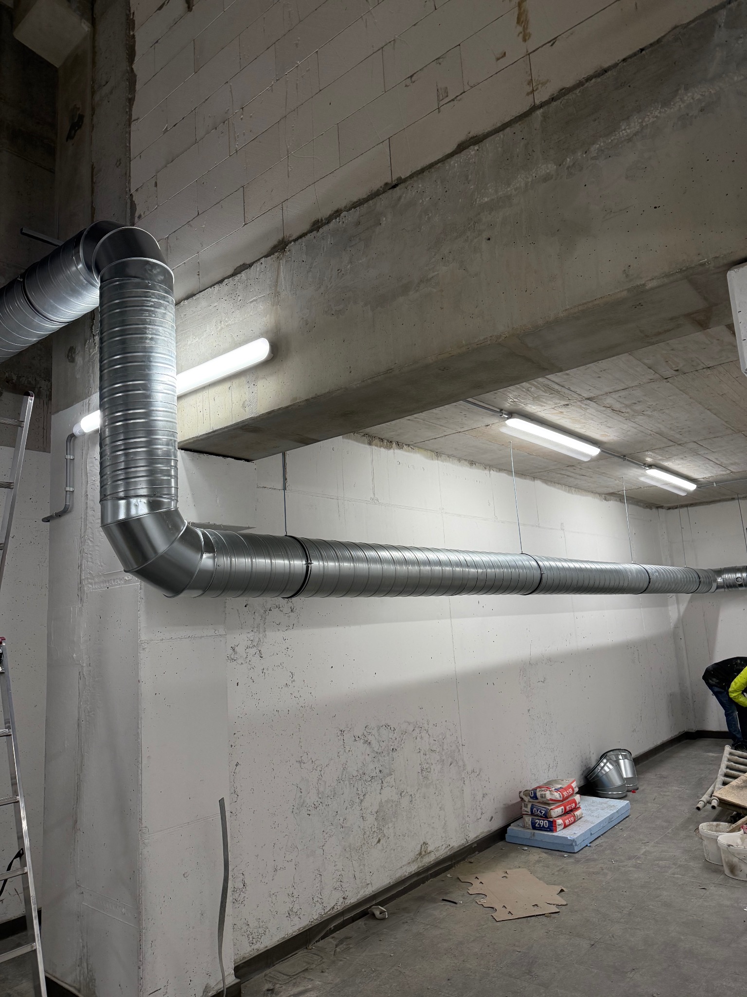 Szare, spiralne rury wentylacyjne zamontowane pod betonowym sufitem w surowym wnętrzu. Widoczne oświetlenie, ściany z bloków i materiały budowlane.