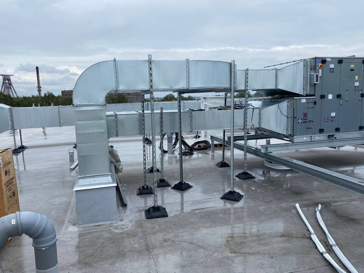 Kompleksowa instalacja wentylacyjna na dachu budynku, z widocznymi kanałami, wspornikami i centralą wentylacyjną LG. Widok na szary, pochmurny dzień.