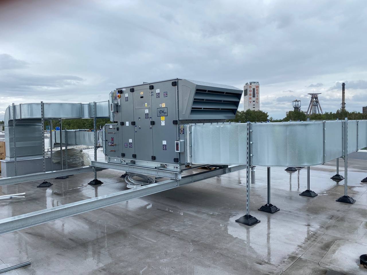 Szara centrala wentylacyjna VBW z kanałami na dachu budynku, widok z niskiej perspektywy, w tle szyby kopalni i napis 'Śląsk'.