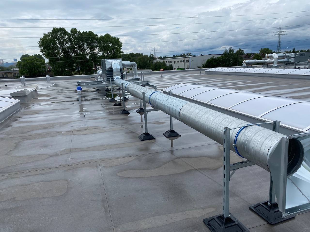 Szare rury wentylacyjne na dachu budynku przemysłowego, wsparte na metalowych stelażach. Widoczne elementy instalacji i tło z zielenią oraz budynkami.