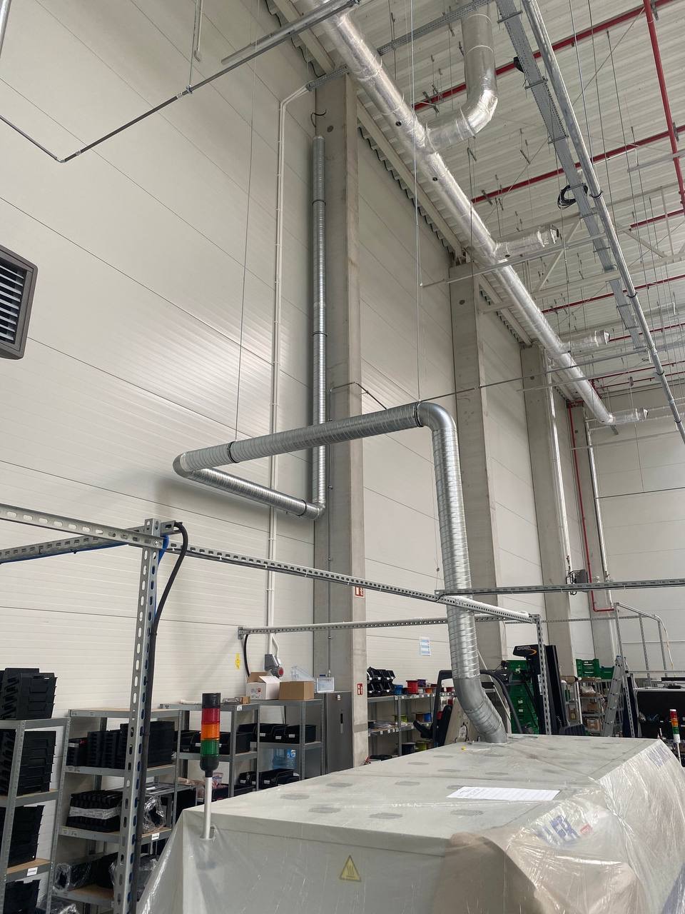 Wnętrze hali produkcyjnej z zamontowaną, srebrną instalacją wentylacyjną pod sufitem. Widoczne rury, elementy mocujące i fragmenty wyposażenia hali.