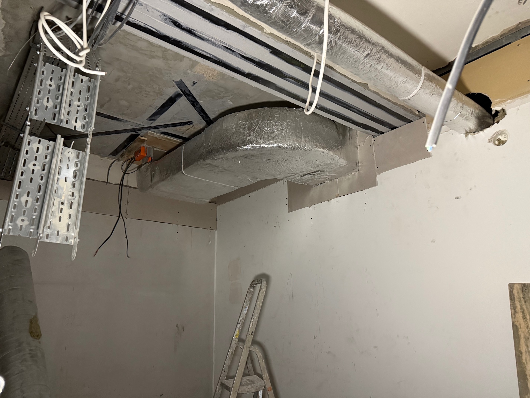 Montaż kanałów wentylacyjnych pod sufitem w surowym wnętrzu. Widoczne przewody, drabina i elementy konstrukcyjne. Etap prac instalacyjnych.