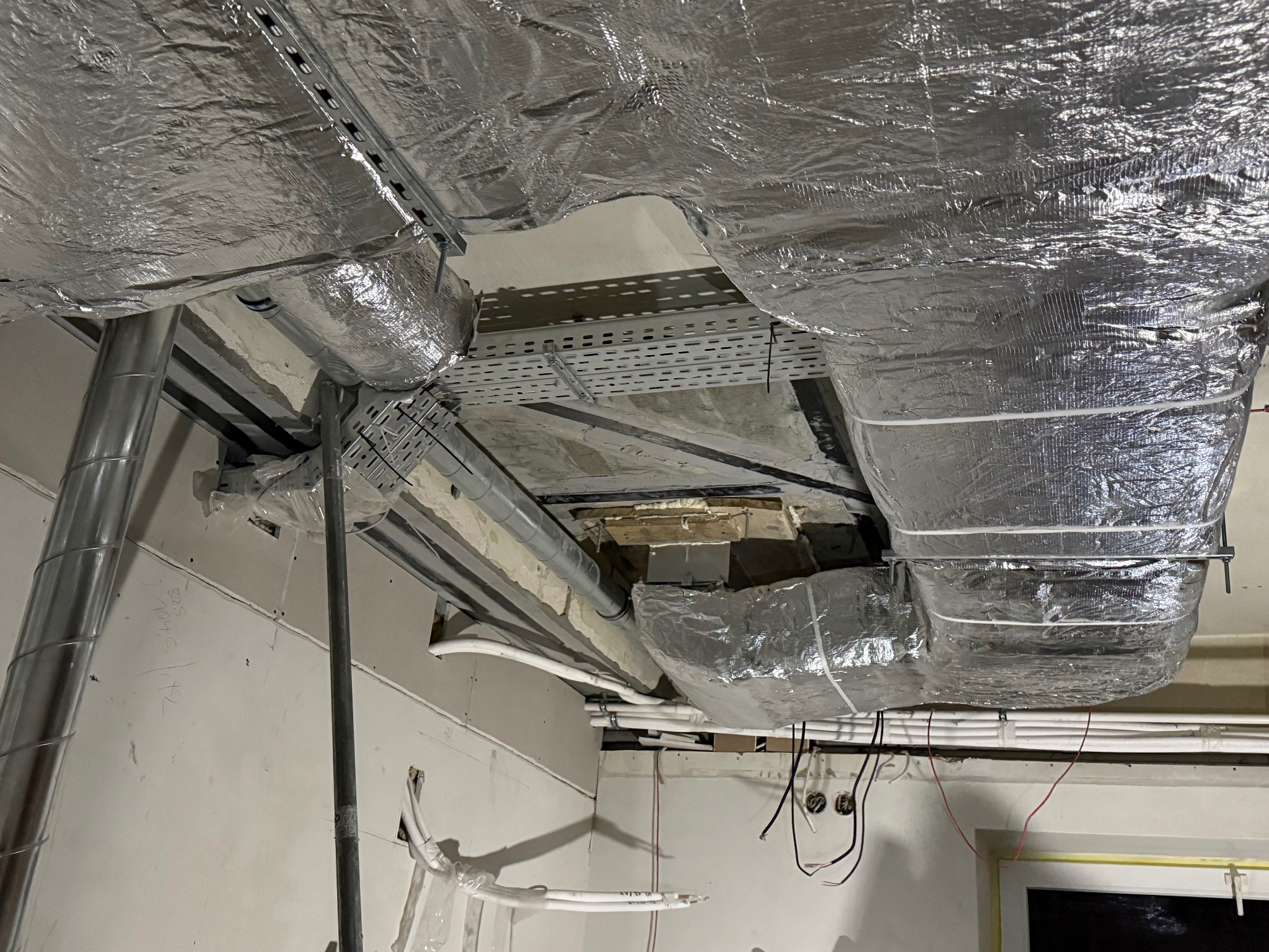 Instalacja przewodów wentylacyjnych z izolacją aluminiową pod sufitem, widoczne koryta kablowe i rury. Prace w toku, surowy stan wykończenia wnętrza.