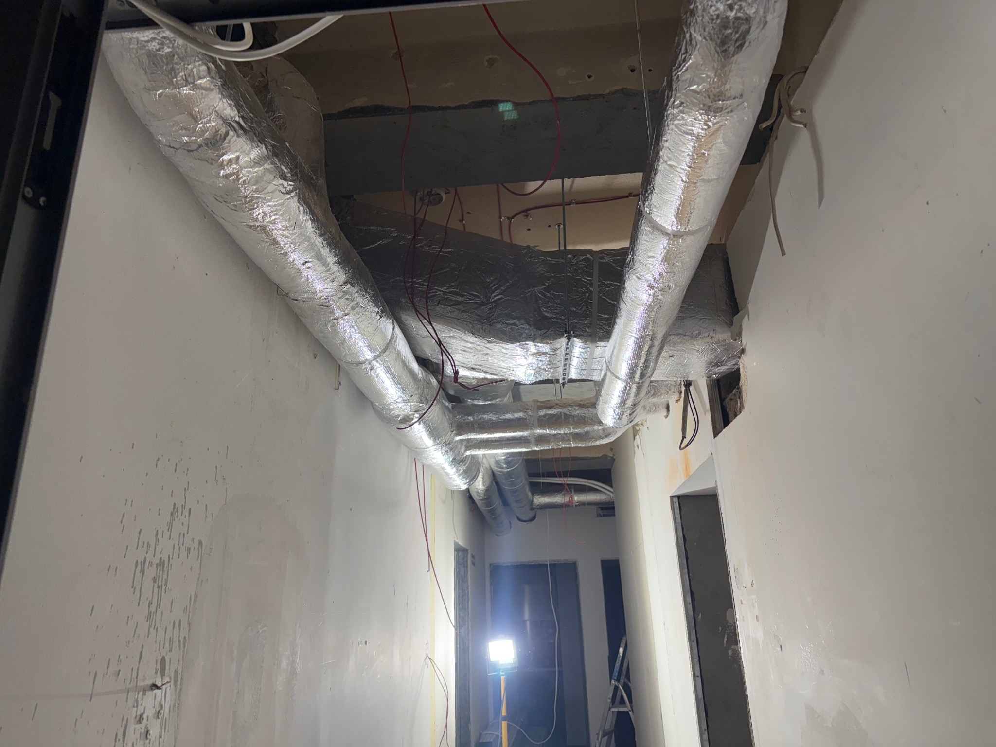 Montaż izolowanych przewodów wentylacyjnych pod sufitem w surowym wnętrzu. Widoczne kable i lampa budowlana. Prace instalacyjne w trakcie realizacji.