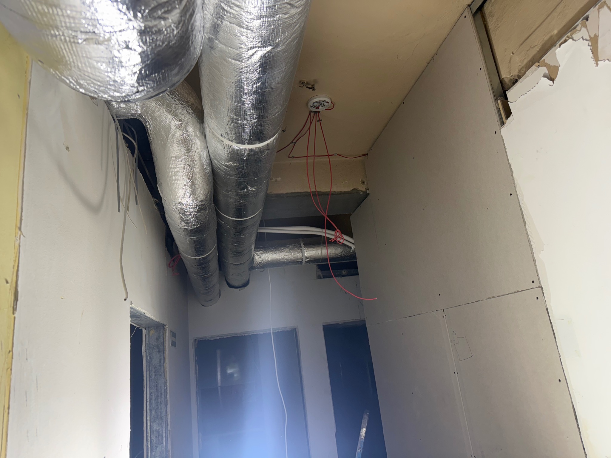 Srebrne rury wentylacyjne pod sufitem w surowym wnętrzu z płyt gipsowych. Widoczne kable i otwory drzwiowe. Etap prac instalacyjnych.